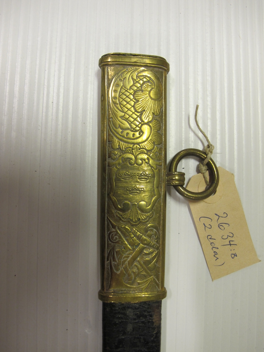 Balja til sjöofficerssabel (O 02634.A) av svartlackerat läder med tre stycken förgyllda och cicilerade beslag. Det översta med lilla riksvapnet, ankare, sponton samt ring. Det mellersta med akterspegel och kampanlykta och ring för koppel. Det understa med drake, spjut, bladverk, ankare, kanon, gevär och hillebard samt nya gösen (efter 1844).