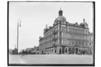 Stenhus i fem våningar med fasad i röd tegel och gul puts. 
Hörnhuset Drottninggatan-Rudbecksgatan.
Bilden har tagits från Våghustorget västerut.