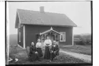 Envånings bostadshus med inredd vind, veranda med snickarglädje.
Fyra personer framför huset.
P. F. Johansson