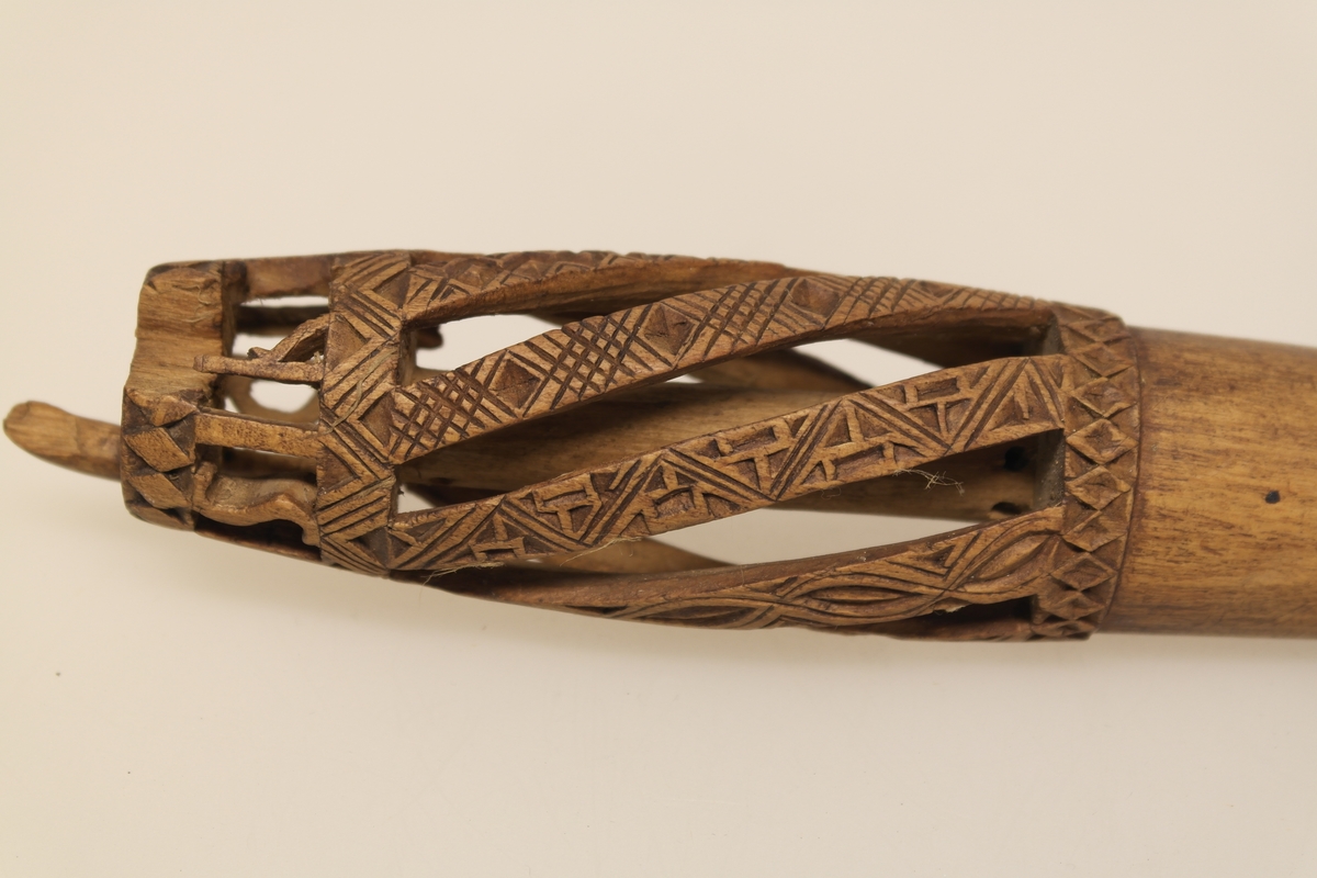 Nøstepinne i skåret furu. Skåret ut med snodde spiler smykket med geometriske mønstre. Øverst på pinnen er det skåret ut, slik at bokstavene står fritt.