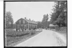 Steninge Slott, nya statbostaden, en och en halvvånings bostadshus, med brutet tak.
Ett uthus till höger på bilden.
Sex män, fem kvinnor och tre barn framför huset. En häst spänd för en vagn.
Uppland