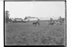 Steninge Slott, stor stensatt ladugård och ekonomibyggnader.
En man med en häst och en kvinna med en tjur framför ladugården.
Uppland