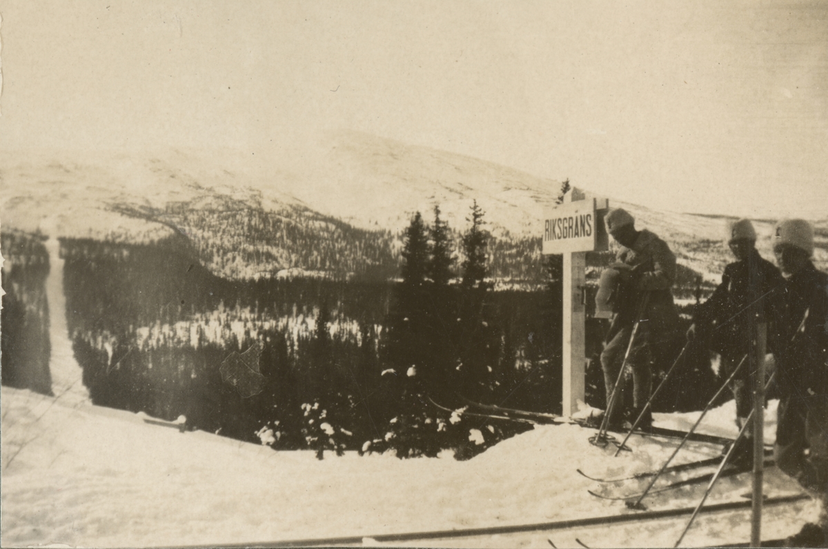 Landsgräns mellan Sverige och Norge. Tre soldater på skidor vid en gränsskylt. Rågången fortsätter genom dalen till berget på motsatta sidan.