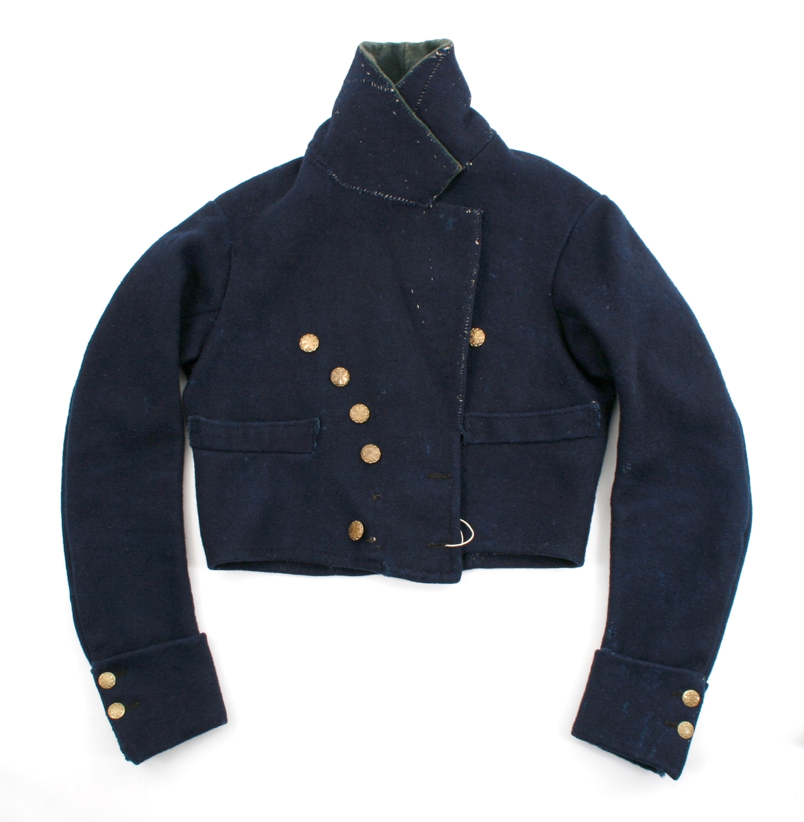 Kort trøye i mørk blått ullstoff, kypertvevd. Overkrage i mørk grønt ullstoff. Underkrage og slagene er mørkeblå. Trøyen er dobbeltspent med buet knappestilling 6 x 2. Den lukkes bare med 2 knapperKnappene er preget med en blomst i midten og en ytterkant av trekanter. På framstykket er det 2lommeklaffer, men bare 1 lomme foret med ubleket linstoff. Kun framstykket er foret. Sidesømmer og skuldersømmer er bakovertrekte. Trange tosømsermer med mansjetter som har 2 messingfargede knapper og 2 knapphull. Stikninger rundt kragen, slagene, MF og nedre kant av trøyen. Høyre undererme er skjøtt oppe ved ermhulen og nede ved mansjetten.