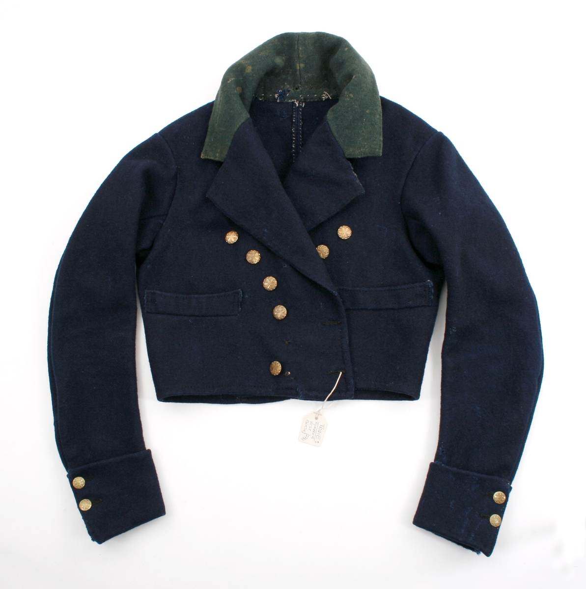 Kort trøye i mørk blått ullstoff, kypertvevd. Overkrage i mørk grønt ullstoff. Underkrage og slagene er mørkeblå. Trøyen er dobbeltspent med buet knappestilling 6 x 2. Den lukkes bare med 2 knapperKnappene er preget med en blomst i midten og en ytterkant av trekanter. På framstykket er det 2lommeklaffer, men bare 1 lomme foret med ubleket linstoff. Kun framstykket er foret. Sidesømmer og skuldersømmer er bakovertrekte. Trange tosømsermer med mansjetter som har 2 messingfargede knapper og 2 knapphull. Stikninger rundt kragen, slagene, MF og nedre kant av trøyen. Høyre undererme er skjøtt oppe ved ermhulen og nede ved mansjetten.