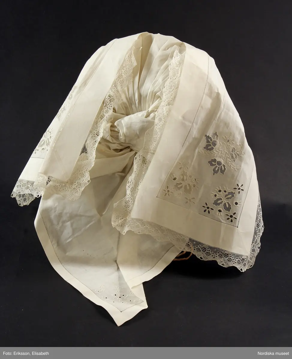 b. Brudtröja av svart vadmal,  Runt alla kanter bård av ytterst brunsvart sammetsband och innanför ett 4 cm brett svart sammetsband  med invävt bladmönster. På ärmarna ett svart slätt sammetsband med pålagd guldspets därefter siden band i rosa och grönt på botten av  svart  sammet och överst i bården en knypplad guldspets. 
c: Förkläde. Material: Textil, bomull. Teknik: Manufakturvävning. Handsömnad. Längd: 77 cm. Bredd: 110 cm. Färg: Vit.
Förkläde i vitt manufakturvävt bomullstyg, ihopsytt i mitten, smalrandigt, stärkt. Rynkningar mot midjelinning i tuskaftat bomullstyg, 1,5 cm bred, fastsydda bomullsband i sidorna. Uppvikt fåll nedtill, 6,5 cm, ovanför fåll, 5 lagda veck med ca 1 cm mellanrum, enkel utdragssöm i kanten av vecken.
/Inga-Lill Eliasson 2008-02-25

e: Krage. Längd: 47,5, cm - totallängd, bröststycke. Bredd: 33 cm - bröststycke. Höjd: 14 cm - kraghöjd, inkl spets. Material: Textil, bomull. Lingarn - spets. Bomullsgarn - broderi. Teknik: Tuskaft. Vitbroderi, Knyppling. Handsömnad.
Krage i tuskaftat vitt bomullstyg. Rektangulär duk, uppklippt halshål med fastsydd krage, 14 cm hög. Kragen har 3,5 cm bred fåll, enkel utdragssöm i kanten. Vitbroderi  i framkant,  plattsöm och kantsydda hål med infällt tyll, två sydda knapphål. Handknypplad spets fastsydd på långsida och korstsidor, 2,5 cm bred. Öppning fram, 24 cm långt sprund. Kragen är handsydd.
Lästips: Svensson Sigfrid, (1935). Skånes Folkdräkter, en dräkthistorisk undersökning 1500 - 1900. Stockholm: Nordiska Museets Handlingar: 3.
/Inga-Lill Eliasson 2008-02-19