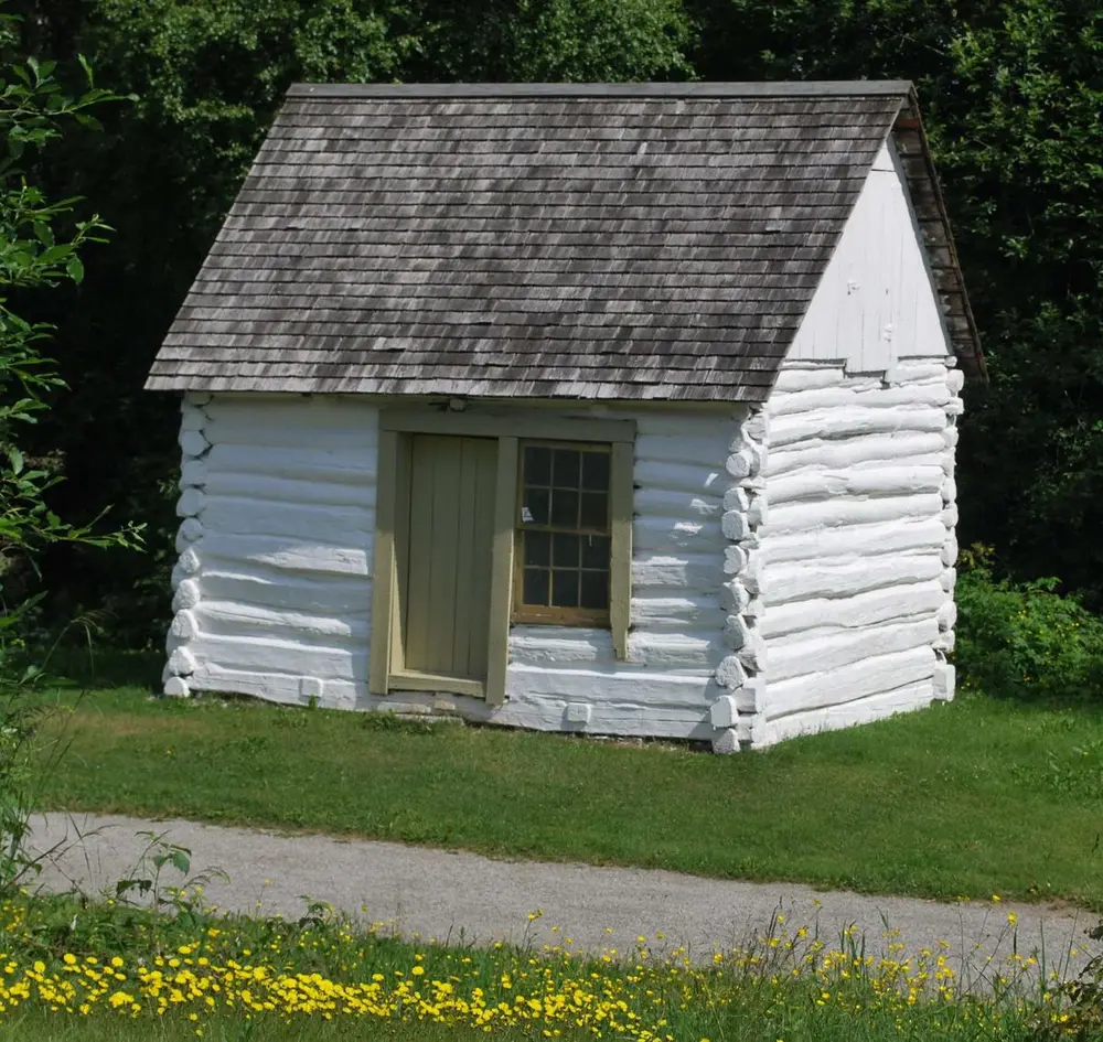 Et lite, en-etasjes, hvitkalket tømmerhus med tre-spon på taket. Gras og blomster på bakken langs en grussti. Trær i bakgrunnen.