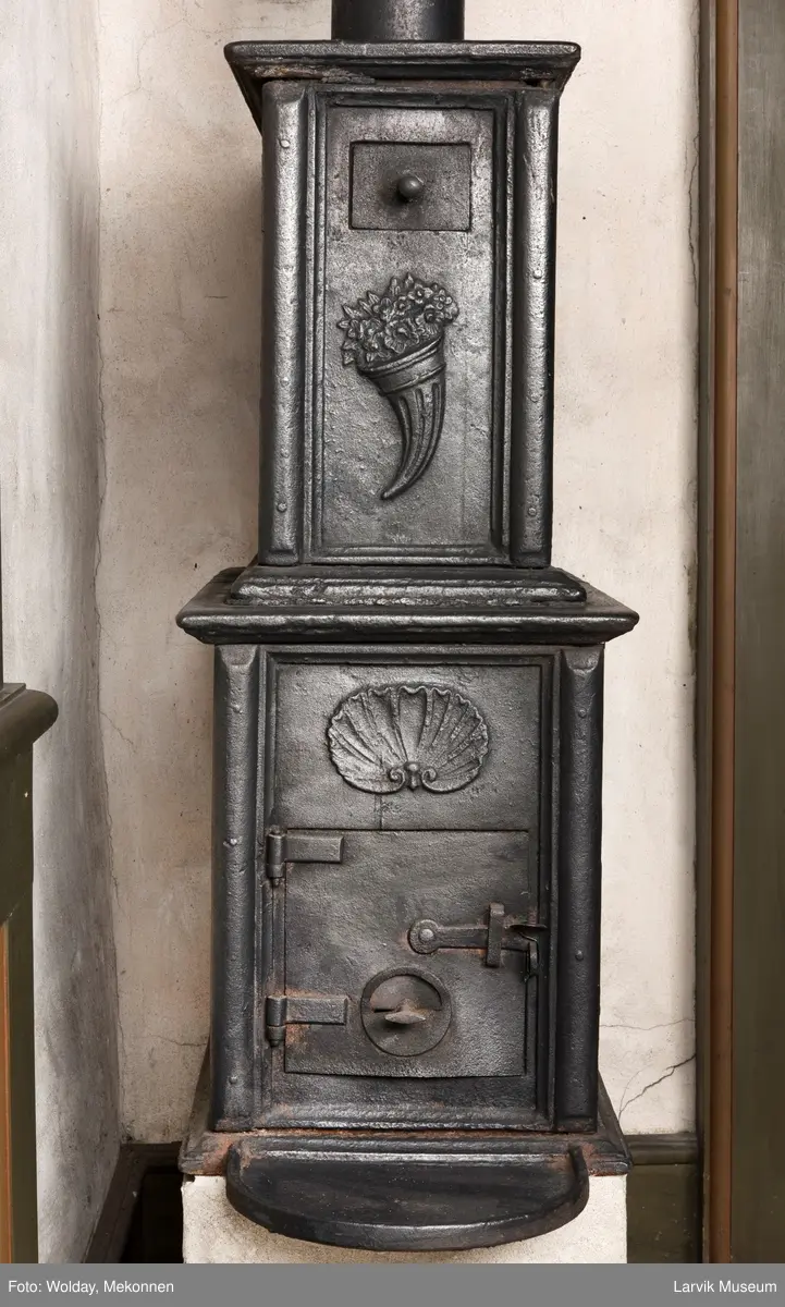 Jernovn med to etasjer, begge er rektangulære. Ovn står på muret fot/understell.
1etasje langside: Vulkan som smed .
1etasje kortside: Musling. 
2etasje langside: Buet åpning i profilert innramming omgitt av girlander. 
2etasje kortside: Overflødighetshorn.