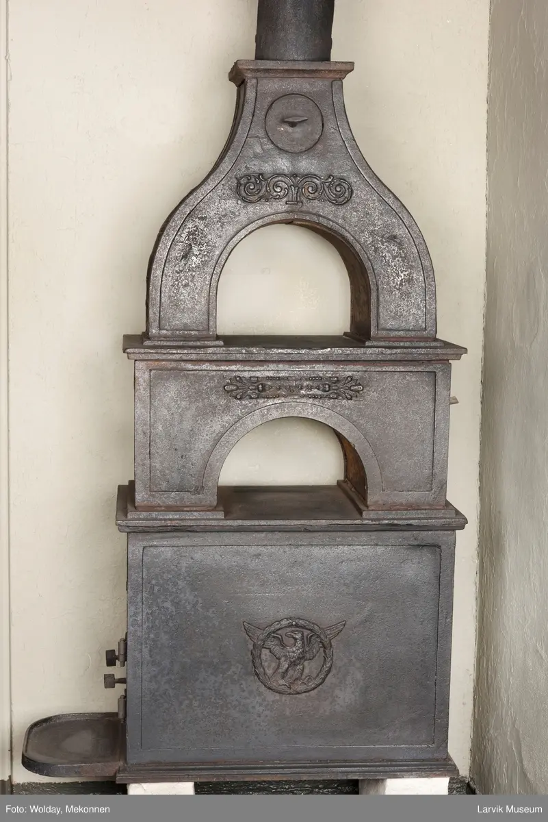 Jernovn i tre etasjer med muret fundament. Langs topp og rundbuet åpning rankeornament. Flatt askebrett med buede hjørner. 
1. etasje langside: Ørn i laurbærkrans 
1. etasje kortside: mrk.: FRITZØE