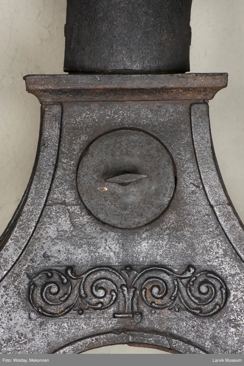 Jernovn i tre etasjer med muret fundament. Langs topp og rundbuet åpning rankeornament. Flatt askebrett med buede hjørner. 
1. etasje langside: Ørn i laurbærkrans 
1. etasje kortside: mrk.: FRITZØE