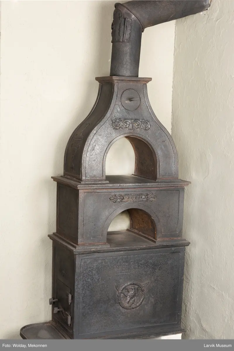 Jernovn i tre etasjer med muret fundament. Langs topp og rundbuet åpning rankeornament. Flatt askebrett med buede hjørner. 
1. etasje langside: Ørn i laurbærkrans 
1. etasje kortside: mrk.: FRITZØE