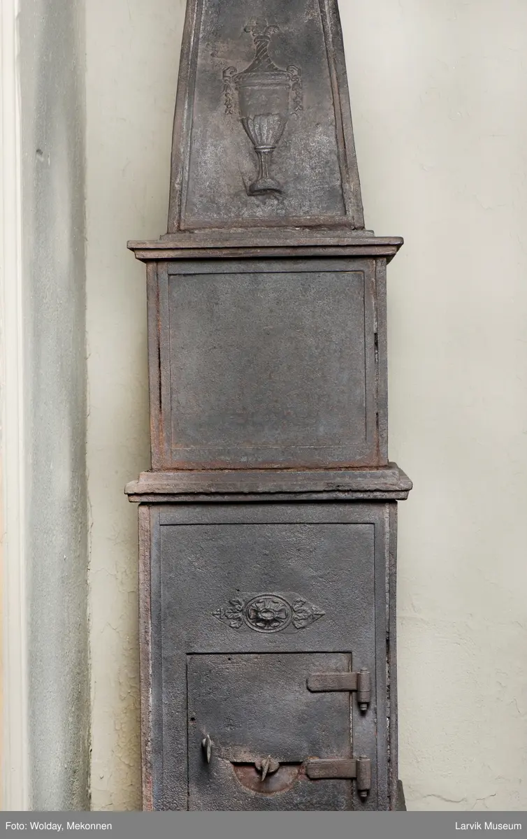 Jernovn i tre etasjer med muret fundament. Langs topp og rundbuet åpning rankeornament. Flatt askebrett med buede hjørner. 
1. etasje langside: Ørn i laurbærkrans 
1. etasje kortside: mrk.: FRITZØE