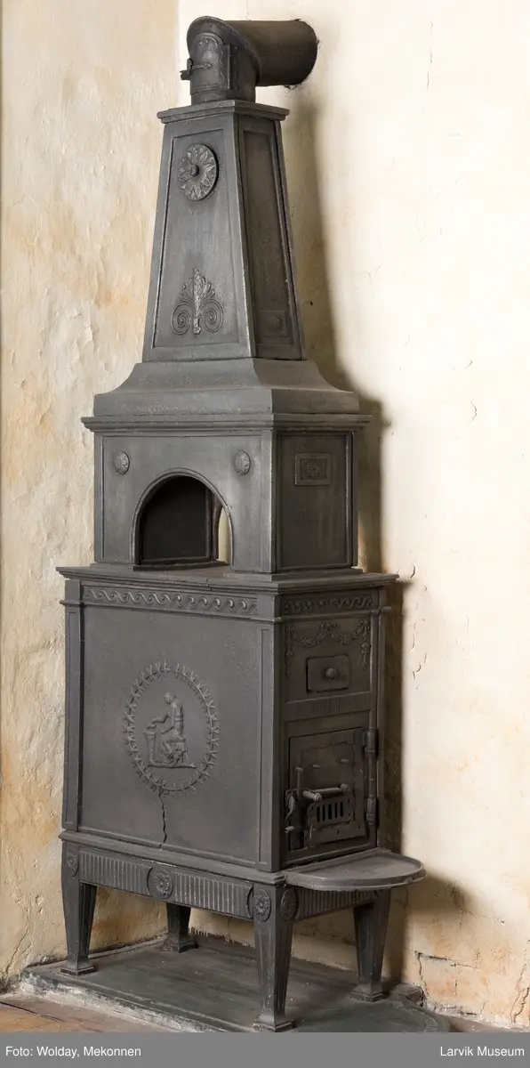 Tredelt , nederste etasje rektangulær fyrkasse/midtf.portalf/pyr.t. ben og sarg med kannelyrer og rosett i tre.  1.etasje langside: bladmedaljong, kvinne ved alter, øverst bølgemeander. 1.etasje kortside: bølgemeander, girlander og rosett 2.etasje langside: rundbuet åpning, i de øvre hjørner 2. etasje kortside: rosett i rektangel topp langside: palmett og rosett topp.Kortside: rosett i rektangel