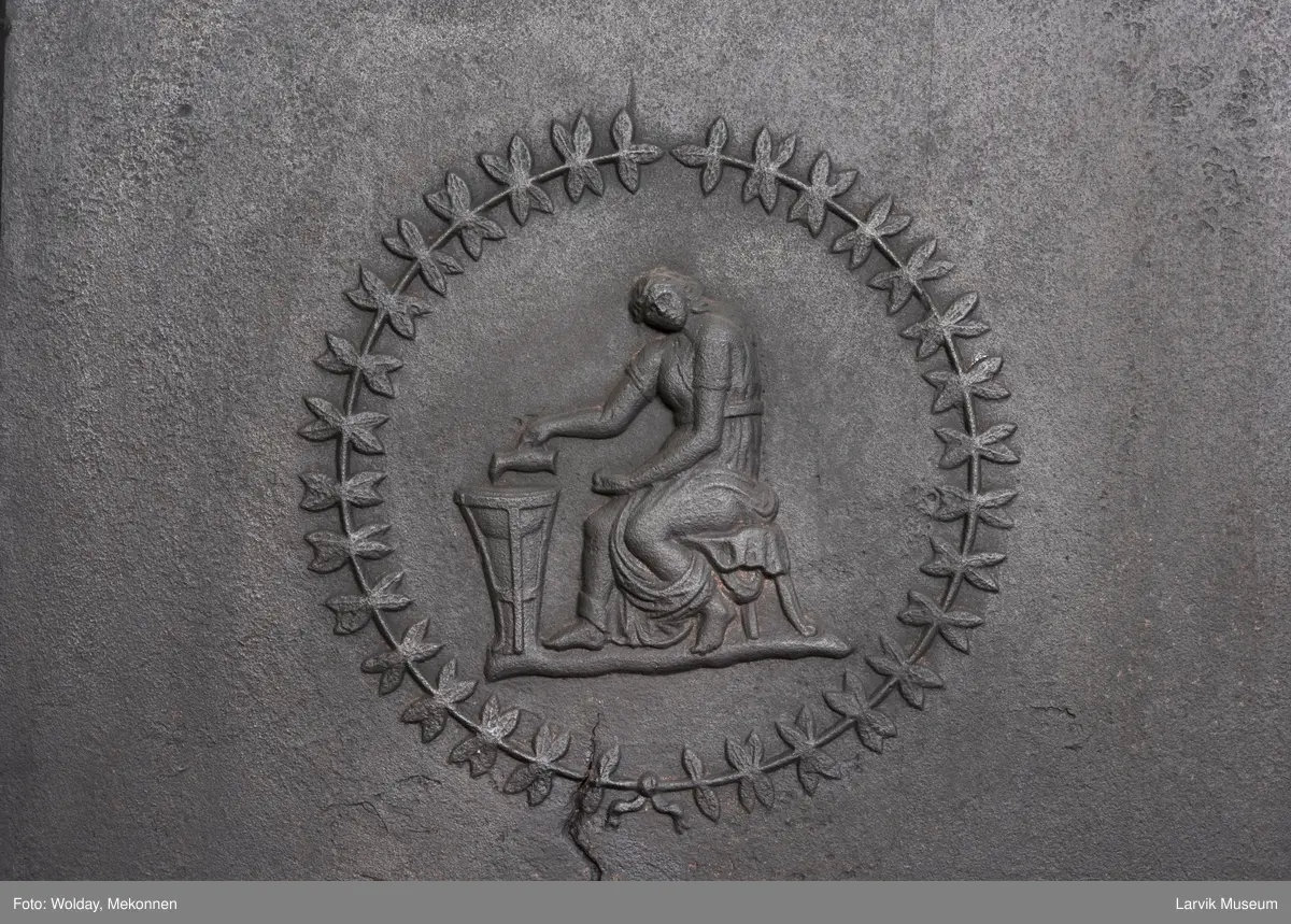 Tredelt , nederste etasje rektangulær fyrkasse/midtf.portalf/pyr.t. ben og sarg med kannelyrer og rosett i tre.  1.etasje langside: bladmedaljong, kvinne ved alter, øverst bølgemeander. 1.etasje kortside: bølgemeander, girlander og rosett 2.etasje langside: rundbuet åpning, i de øvre hjørner 2. etasje kortside: rosett i rektangel topp langside: palmett og rosett topp.Kortside: rosett i rektangel