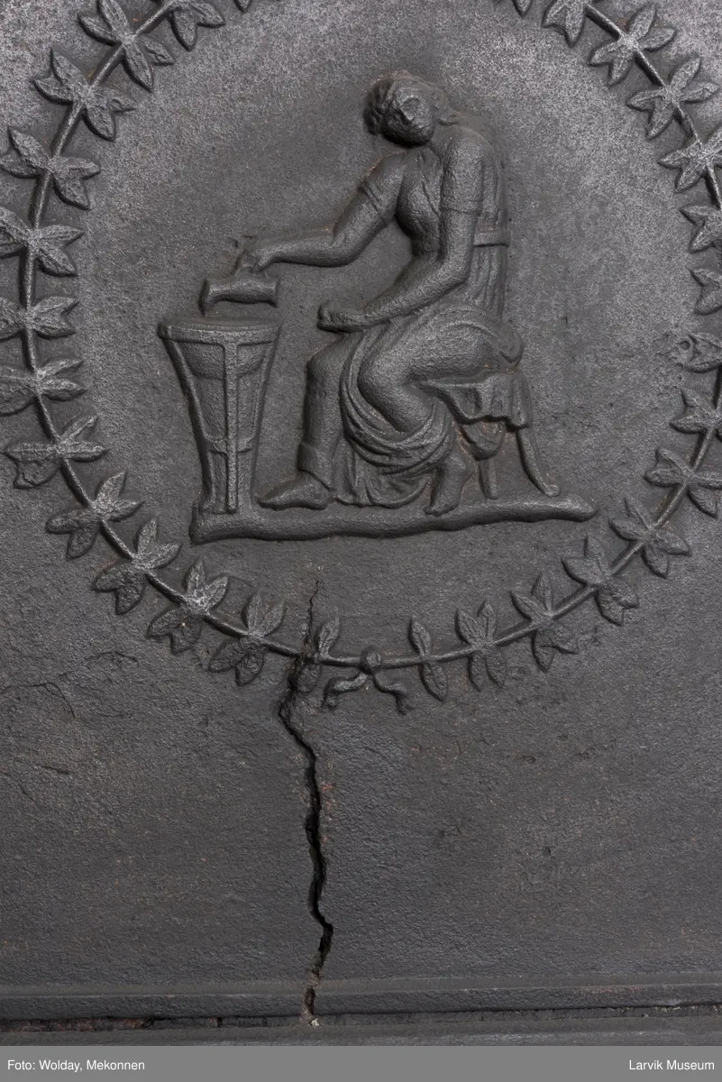 Tredelt , nederste etasje rektangulær fyrkasse/midtf.portalf/pyr.t. ben og sarg med kannelyrer og rosett i tre.  1.etasje langside: bladmedaljong, kvinne ved alter, øverst bølgemeander. 1.etasje kortside: bølgemeander, girlander og rosett 2.etasje langside: rundbuet åpning, i de øvre hjørner 2. etasje kortside: rosett i rektangel topp langside: palmett og rosett topp.Kortside: rosett i rektangel