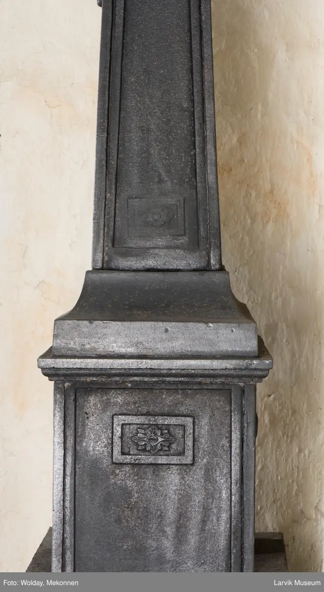 Tredelt , nederste etasje rektangulær fyrkasse/midtf.portalf/pyr.t. ben og sarg med kannelyrer og rosett i tre.  1.etasje langside: bladmedaljong, kvinne ved alter, øverst bølgemeander. 1.etasje kortside: bølgemeander, girlander og rosett 2.etasje langside: rundbuet åpning, i de øvre hjørner 2. etasje kortside: rosett i rektangel topp langside: palmett og rosett topp.Kortside: rosett i rektangel