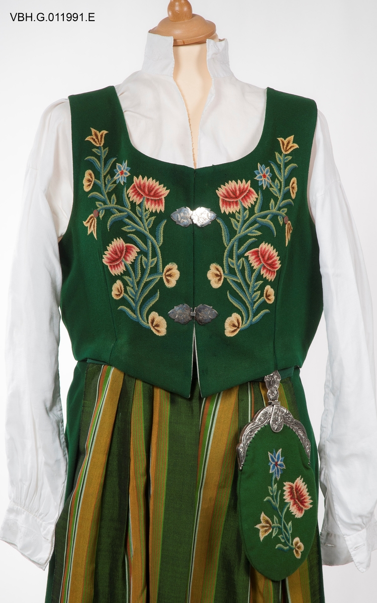 Broderier med blomstermotiver. Sjal og forkle med rutemønster.
Kjolen har blitt sydd ut i senere år med grønt bakstykke på 7 cm på hver side.