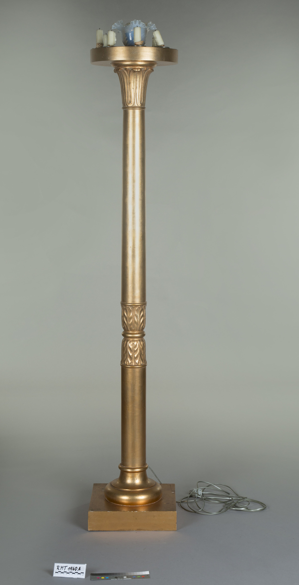 Forgylte gulvstående lamper for flere lys, både stearinlys og elektrisk. Hull til stearinlys langs kanten på hele topp-platen. En åpen lampeskjerm i glass omkranser det elektriske lyset midt på abakus, med bølgende kant (som en fakkel). Lampen er formet som en frittstående gresk søyle med base. Glatt utforming, med et område med bladutskjæringer like under midten, samt på kapitélet. Rund abakus og firkantet base.