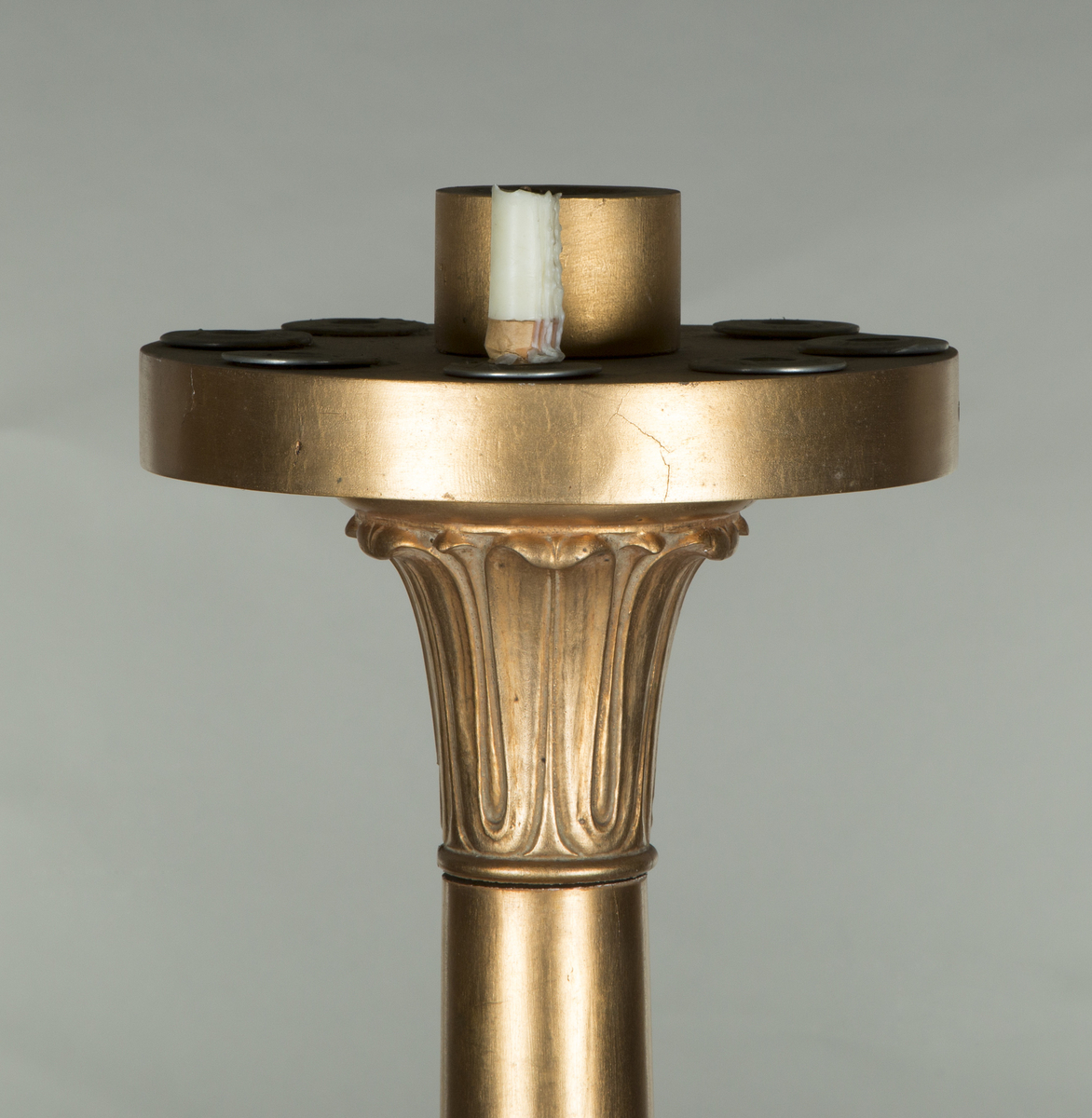 Forgylte gulvstående lamper for flere lys, både stearinlys og elektrisk. Hull til stearinlys langs kanten på hele topp-platen. En åpen lampeskjerm i glass omkranser det elektriske lyset midt på abakus, med bølgende kant (som en fakkel). Lampen er formet som en frittstående gresk søyle med base. Glatt utforming, med et område med bladutskjæringer like under midten, samt på kapitélet. Rund abakus og firkantet base.