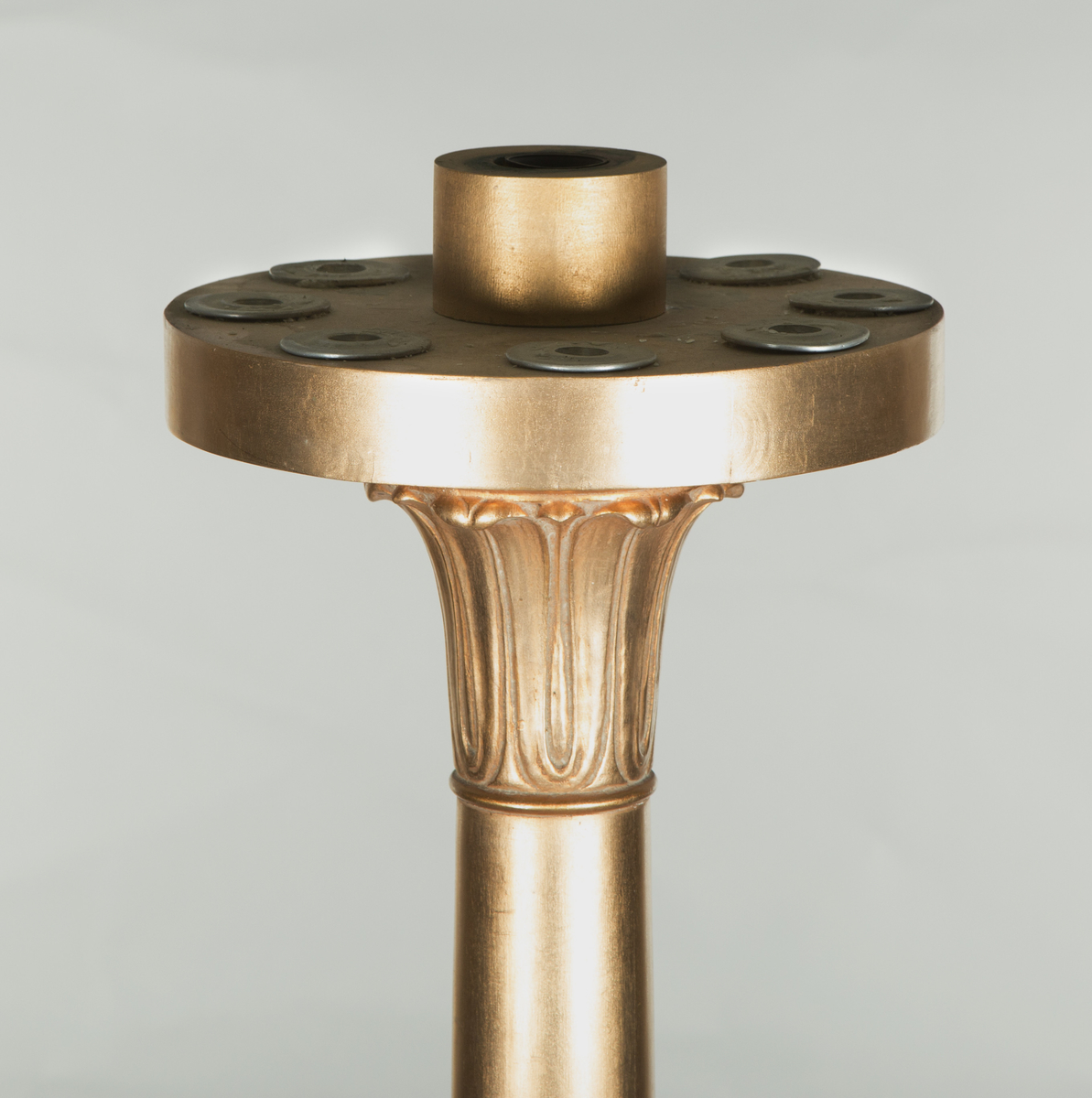 Forgylte gulvstående lamper for flere lys, både stearinlys og elektrisk. Hull til stearinlys langs kanten på hele topp-platen. En åpen lampeskjerm i glass omkranser det elektriske lyset midt på abakus, med bølgende kant (som en fakkel). Lampen er formet som en frittstående gresk søyle med base. Glatt utforming, med et område med bladutskjæringer like under midten, samt på kapitélet. Rund abakus og firkantet base. 
To av lampene har et forgylt tresylinder rundt pipen for det elektriske lyset. 
Var sannsynligvis ikke el.belysning originalt.