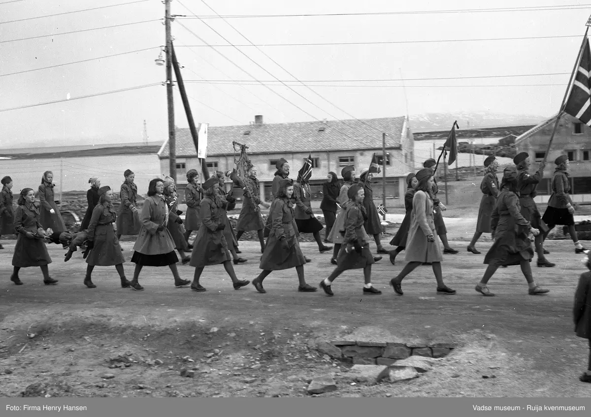 Vadsø sentrum 1951. Del av 17.maitog i Hvistendalsgate. Bildet viser ca. 20 jenter, noen i uniform og alle med speiderlue fotografert fra nord. I bakgrunnen sees Luftskipsmasta på Vadsøya og forretningsbygg under bygging: Torleif Mathisen og Leif Bjerck.
