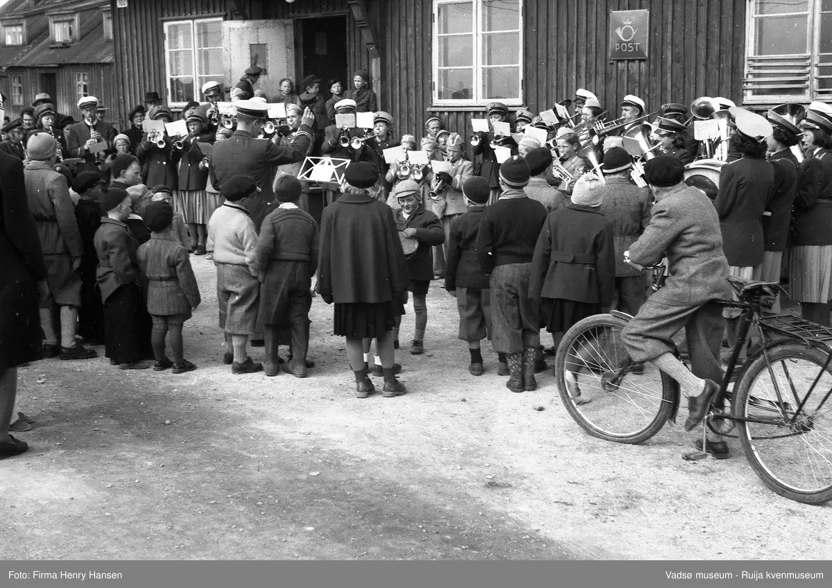 Vadsø sentrum 17. mai 1951, Musikkorps står oppstilt og spiller, jenter, gutter og voksne, med dirigent. Aksel Niska er helt til høyre i korpset. Flere barn står foran korpset med ryggen til, en av guttene har sykkel. Korpset står foran postbrakka med skilt "Post "og nr "102" på veggen. Vi ser litt av nabohuset til venstre, også en brakke.