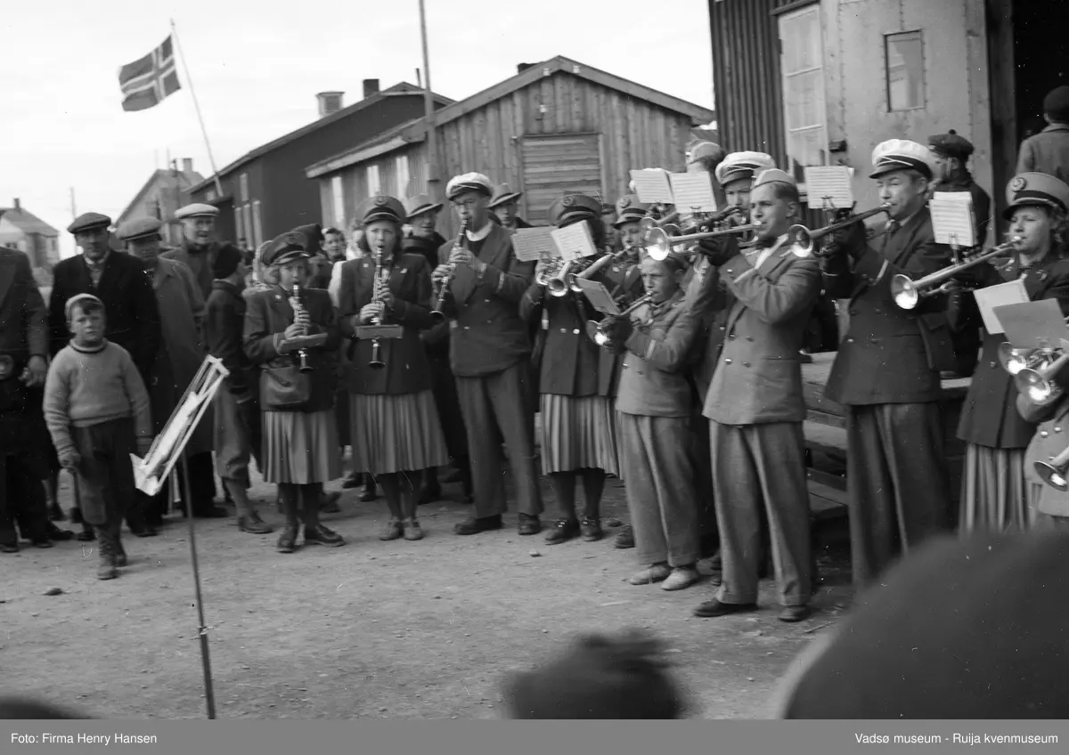 Vadsø sentrum 17. mai 1951, Musikkorps står oppstilt og spiller foran postbrakka. Voksne og barn er tilskuere. Vi ser deler av fire andre bygninger, to bolighus og en brakke.