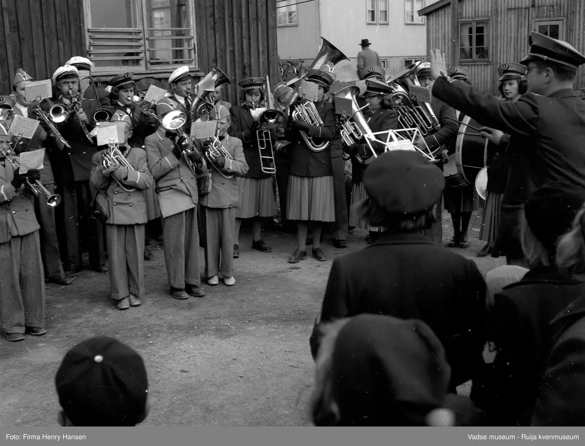 Syttende mai i Vadsø 1951. Vadsø jentekorps og Guttemusikken spiller, de står oppstilt mellom brakker. De er kledd i uniformer og spiller etter noter.  Dirigenten Olav Holmen i forgrunnen og Aksel Niska spiller ventilbasun i front. Andre personer som vi ser på bildet er bl.a.: Anne Marie Berg (frisør, gift Lamo), Ingrid Iversen og Liv Danielsen (gift Dallavara).