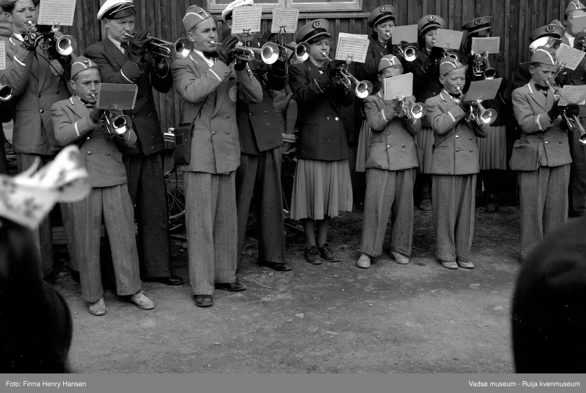 Vadsø sentrum 17. mai 1951. Musikkorps i uniformer, bestående av barn og voksne, står oppstilt og spiller. Vi ser en del av korpset, ikke dirigenten.  Korpset står foran postbrakka.