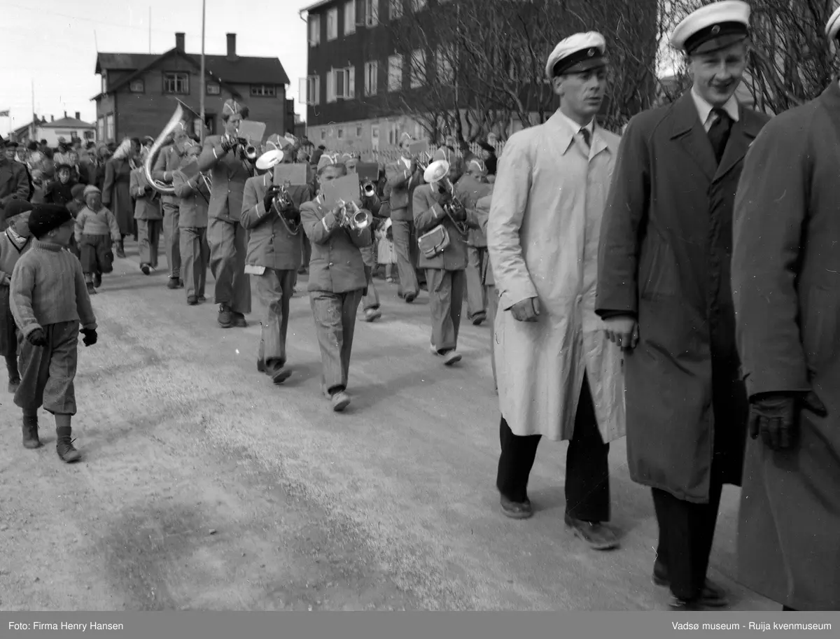 Vadsø 17. mai 1951. Musikkorps marsjerer i gaten. Vi er i Indrebyen i Nyborgveien og "Alders Hvile" er til høyre i bildet. Vadsø Mannsangforening med sangerluer marsjerer i forkant av bildet. Mannssanger i lys frakk er Hans Dagfinn Iversen. Publikum, barn og voksne marsjerer ved siden av og etter korpset.