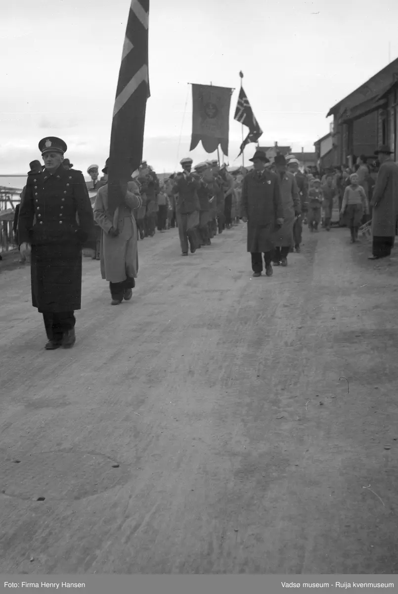 Vadsø 17. mai 1951. Musikkorps masjerer i Indrebyen i Havnegata. Vi ser flagg og fane. Fremst i toget masjerer Wickstrøm(politi). Publikum, barn og voksne sees langs 17.maitoget. I høyre billedkant skimter vi fjorden med kaier.