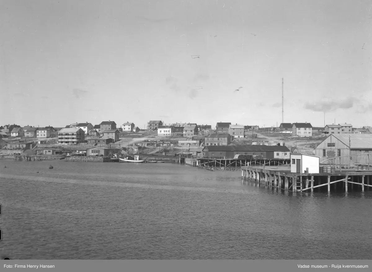Vadsø 1948 med deler av havnen, sentrum og indrebyen. Gjenreisningen er i full gang. Vi ser kaianlegg med bygninger og fiskebåter. Vi ser også ruiner etter kaianelgg og branntomter etter bygninger. Til høyre i bildet ser vi Tollbugata som går fra sentrum og opp mot Damsveien. Til høyre sees en av radiomastene.