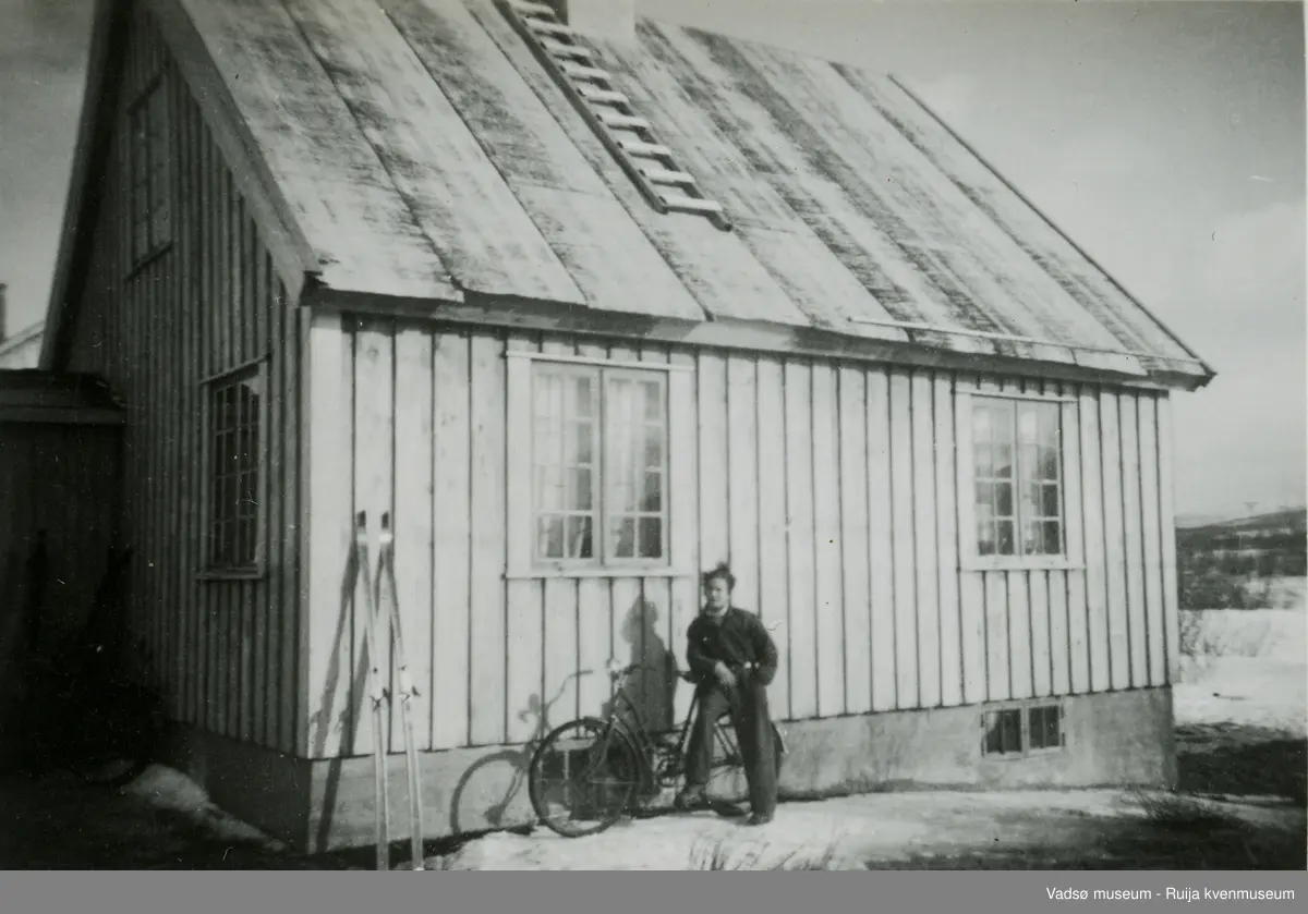 Vadsø. Bildet viser et bolighus med en og en halv etasje, fotografert nordover. Det er trolig et gjenreisningshus, en og en halv etasje. Det er to vinduer mot sør, ett i første etasje og ett i andre etasje mot vest og gardiner i vinduene. Inngangspartiet er mot nordvest. Foran huset sitter en mann på en damesykkel, et par ski står mot veggen. Litt snø ligger på bakken, så bildet er trolig tatt på vårparten.
