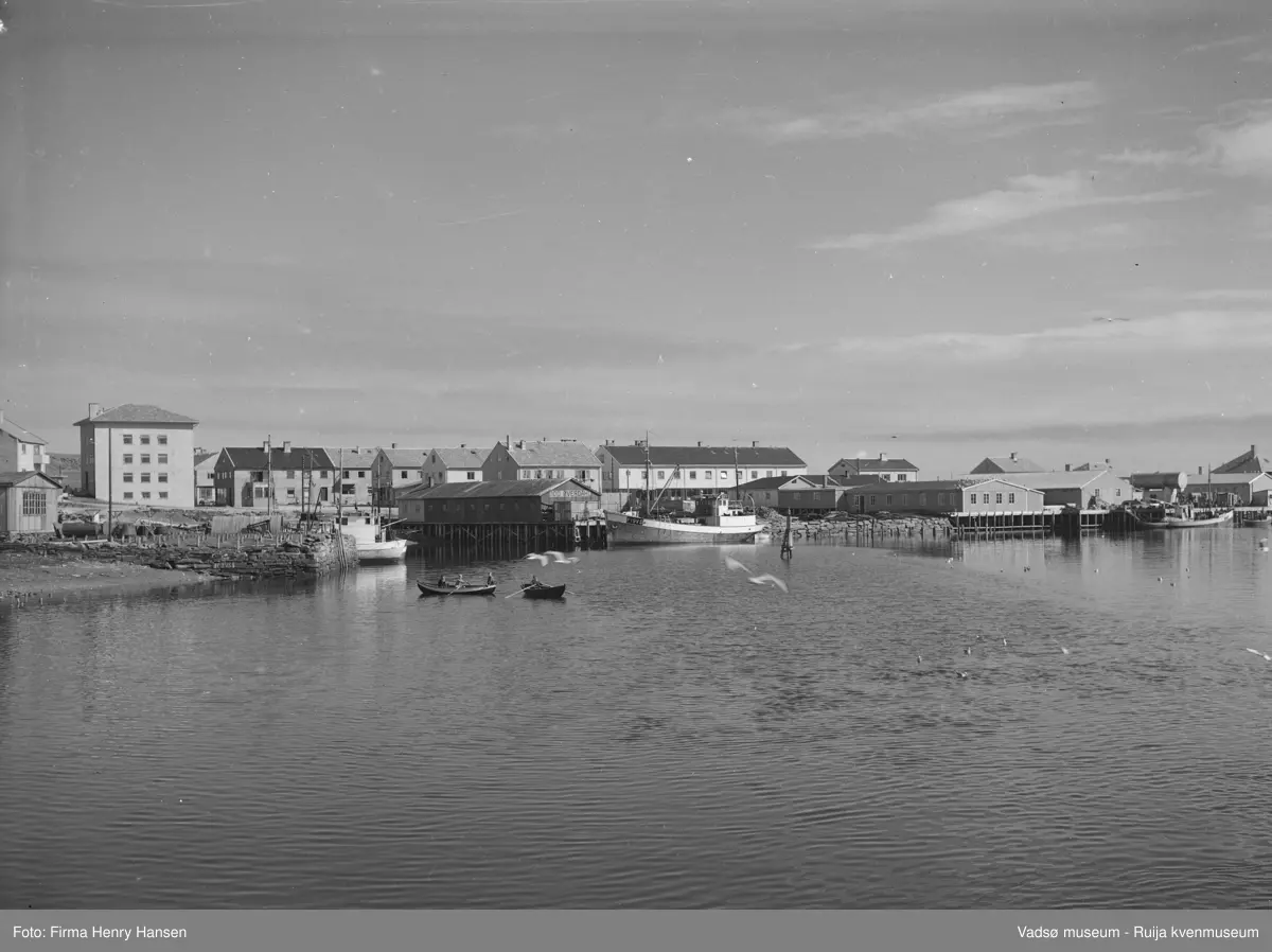 Vadsø Sentrum på 1950-tallet. Vi ser Rådhuset. Torget omkranses av forretningsbyggene til Baker Henriksen, Henry Hansen og Rolf Hansen. Vi ser også forretningsbygget til H.F.Esbensen. På torget kan skimtes barn på sykler. Fremst i bildet ser vi deler av indre havn med måser og to robåter med folk i. Vi ser også flere fiskebåter og kaier med kaihus. Ett av kaihusene har skiltet; Odd Øverdahl.