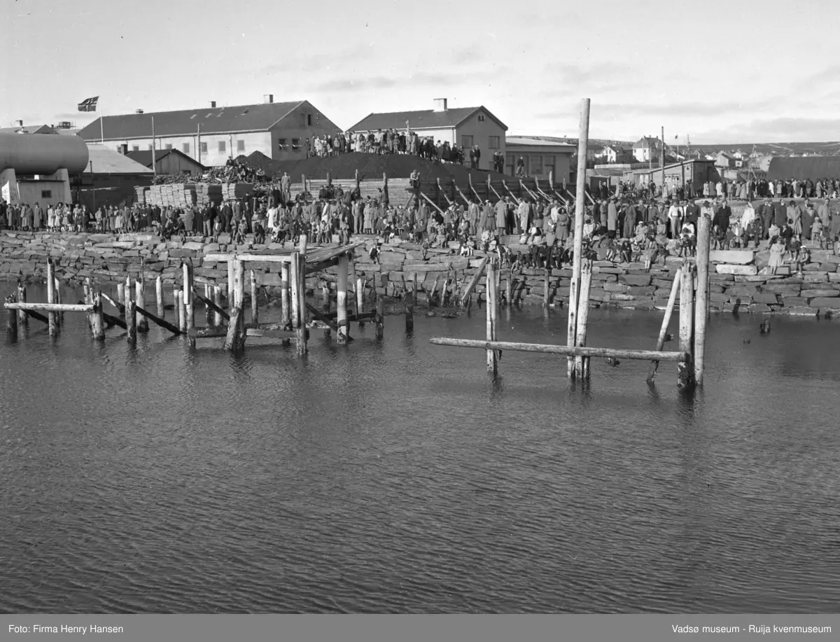 Kongebesøk i Vadsø i 1946. Bildet er tatt mot nordøst. Vi ser forretningsbygg og bygningen til Aust-Finnmark Meieri. I bakkant ser vi Kysthospitalet. Vi kan se en stor folkemengde langs havna. Folk er pyntet, og flagget vaier i vinden. Folk sitter og står og vi ser også noen mennesker noen som står oppå et stort lager av kull. I forkant av bildet ser vi er havna med rester av kaier og kaistolper. I venstre billedkant deler av en stor oljetank. Midt i bildet er H.F. Esbensens butikk.
forretningsbygg og bygningen til Aust-Finnmark Meieri. I bakkant ser vi Kysthospitalet.