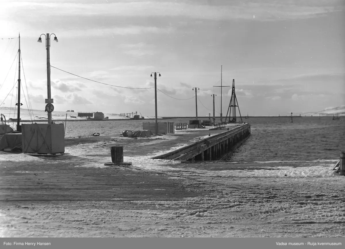 Vadsø april 1952. Bildet er tatt fra dampskipskaien fra øst mot vest. Vi ser kaien og kaispissen med lysstolper, oljefat, et skilt med parkering forbudt/sykling forbudt og landgangen til hurtigruta. Til venstre i bildet ser vi Vadsøya og kaianlegg med kaihus, nothjul og hjellbruk bl.a. med fiskegarn til tørk. Lengst bort i bildet, mot vest, ser vi moloen som går fra Vadsøya mot nord og et par fiskebåter på havna.