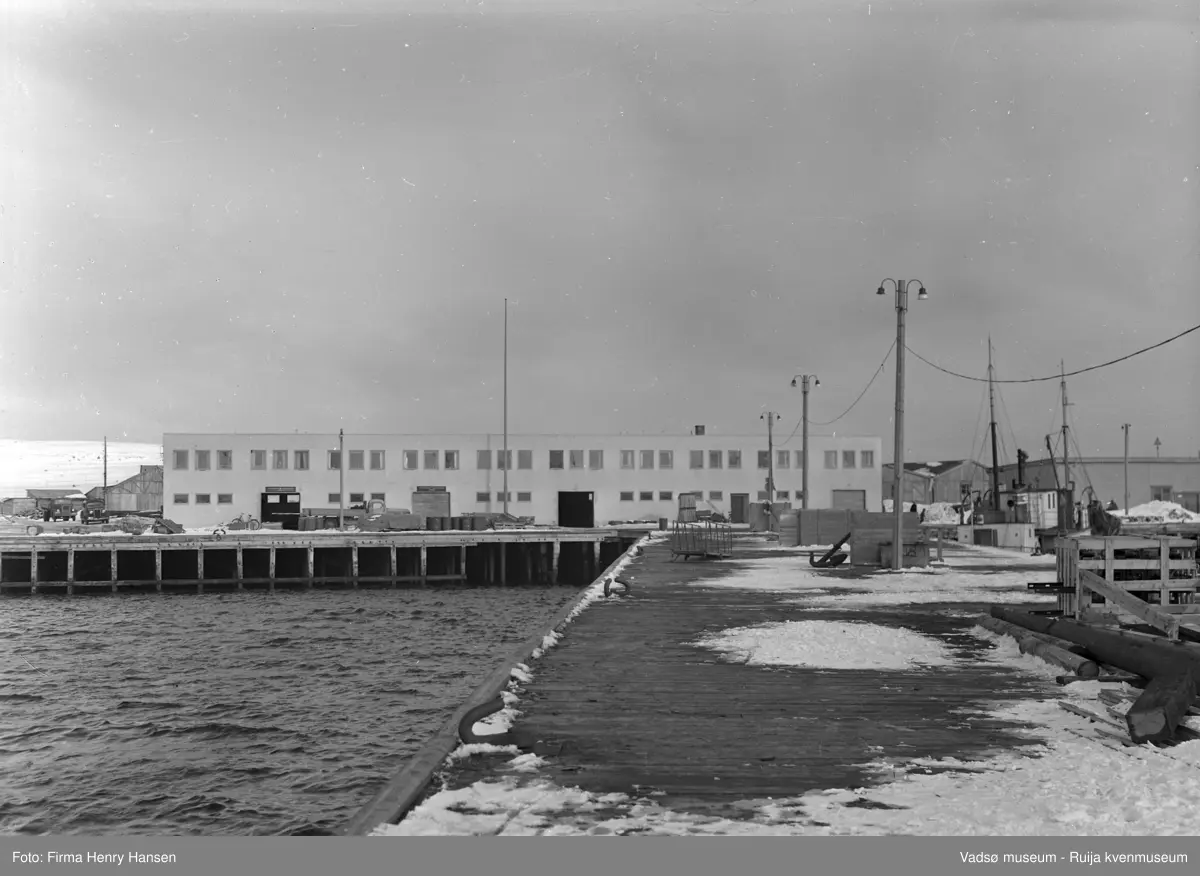 Vadsø april 1952. Bildet er tatt fra spissen av dampskiskaia fra vest mot øst. I forkant av bildet ser vi kaia med snøflekker, lysstolper, et anker og landgangen til hurtigruta. I høyre billedkant fiskebåter og et par bygninger. I bakkant og midt i bildet dampskipsekspedisjonen hvor vi bl.a. ser en lang mast/stolpe, oljetønner, sykler, og en lastebil.
I venstre billedkant ses et par brakker og et par biler.