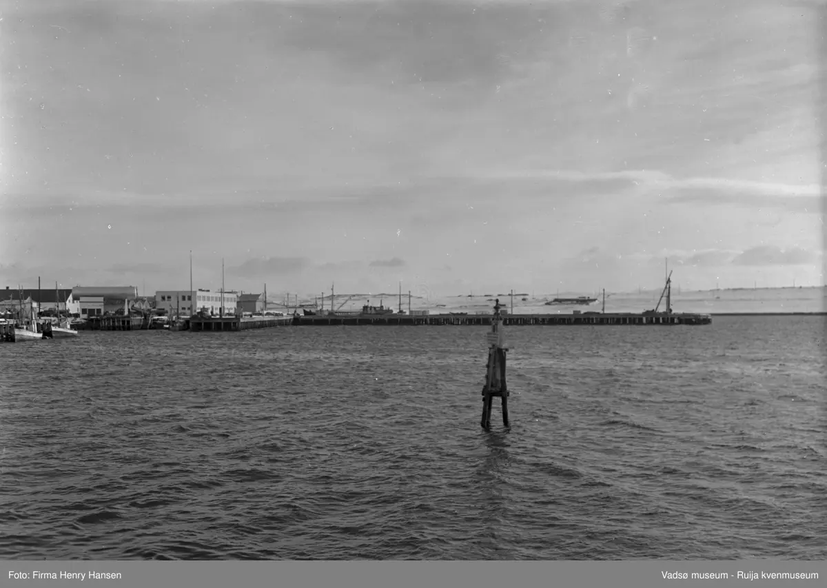 Vadsø april 1952. Bildet er tatt fra fastlandet mot syd. Vi ser havna med Dampskipskaia og dampskipsekspedisjonen midt i bildet. Videre ser vi flere kaianlegg, fiskebåter, tanker, bygninger. I bakre billedkant ser vi Vadsøya og noen bygninger.