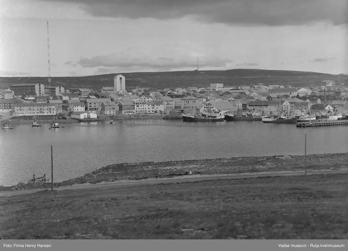 Motivet er Vadsø sentrum sett fra Vadsøya vest for broen 20.07.1968. Øverst ser en Fylkesbygget og Statens hus, Prestegården og Vadsø kirke. Det store bygget øverst til venstre er Vadsø kretsfengsel. Andre offentlige bygg er Vadsø rådhus, Vadsø skole og Vadsø sykehus. Til venstre på havna sees små fiskebåter. Ved kaiene mellom det hvite huset i midten av bildet som er Hermetikkfabrikken og den ytterste delen av dampskipskaia ser en flere større fiskefartøy. I forgrunnen ser man en del av veien som går fra brua til Sildoljefabrikken på Vadsøya.