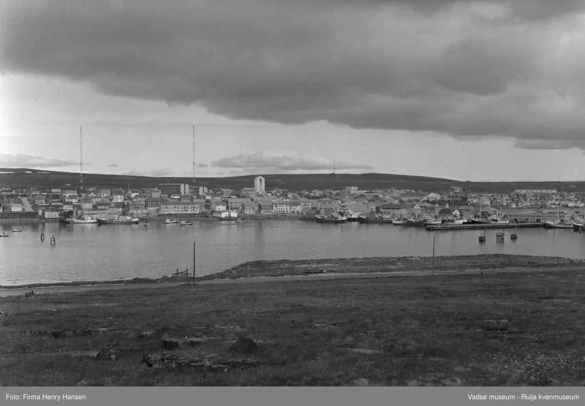 Motivet er Vadsø sentrum sett fra Vadsøya vest for brua 20.07.1968. Øverst ser en Fylkesbygget og Statens hus mellom de to radiomastene, videre mot øst Prestegården og Vadsø kirke. Andre offentlige bygg som sees er Vadsø rådhus, Vadsø skole,  Vadsø sykehus og Vadsø kretsfengsel et stykke ovenfor sykehuset.  Flere store og små fiskebåter ses på havna og dampskipskaia med havneterminalen til høyre. I forgrunnen sees en del av veien som går fra brua til Sildoljefabrikken på Vadsøya.