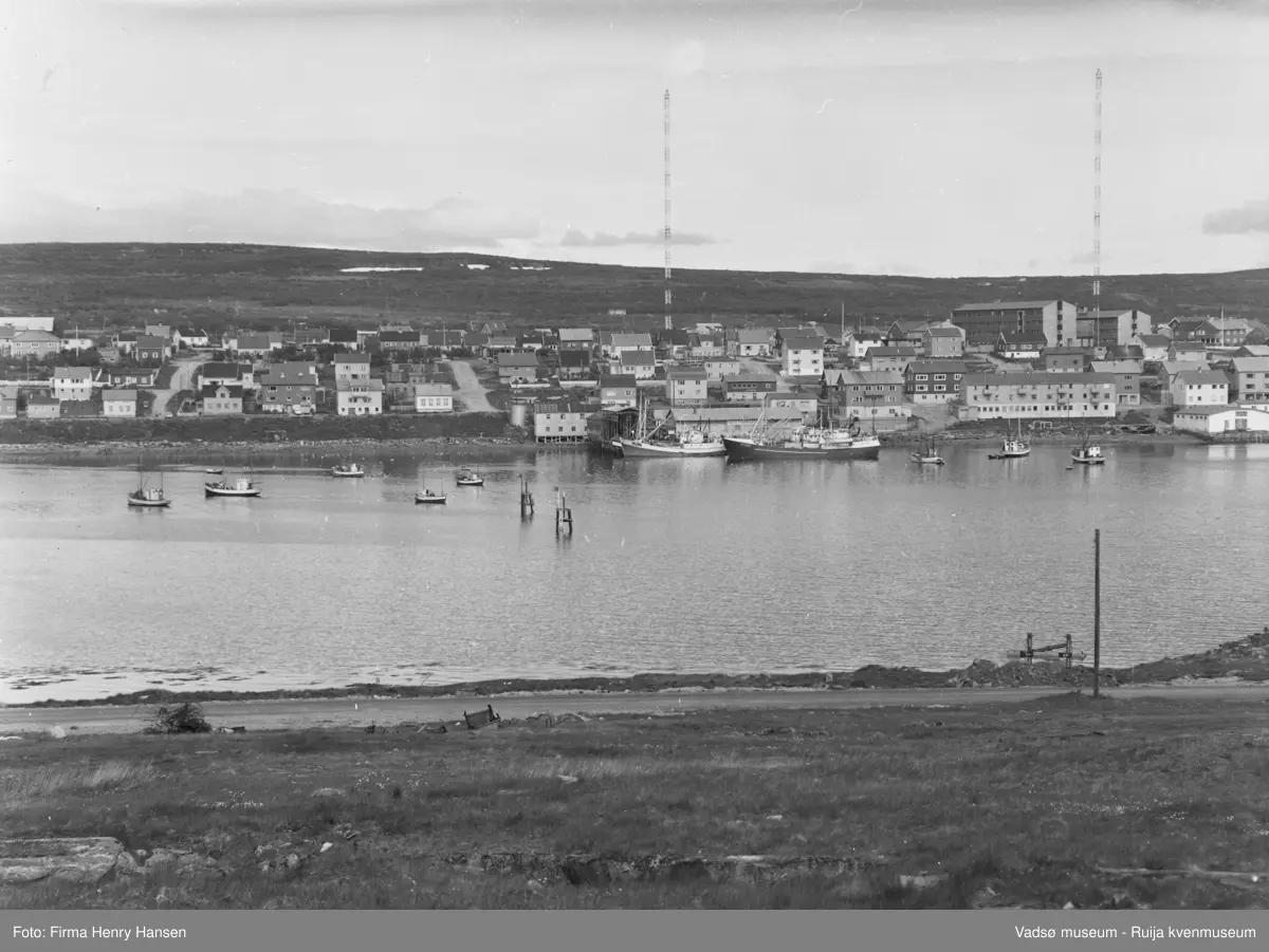 Motivet er vestre del av Vadsø sentrum og havn sett fra Vadsøya vest for Tollbugata 20.07.1968. Øverst ser en de to radiomastene, og til høyre Fylkesbygget, Statens hus og prestegården. På havna sees store og små fiskebåter. Havnegata og parallellgatene Nyborgveien og Damsveien går langs hele bildet. Til venstre i bildet ser en bolighus i disse gatene og mot høyre i Havnegata, forretningsbygg og kaianlegg.  I forgrunnen ser en en del av veien som går fra brua til Sildoljefabrikken på Vadsøya.