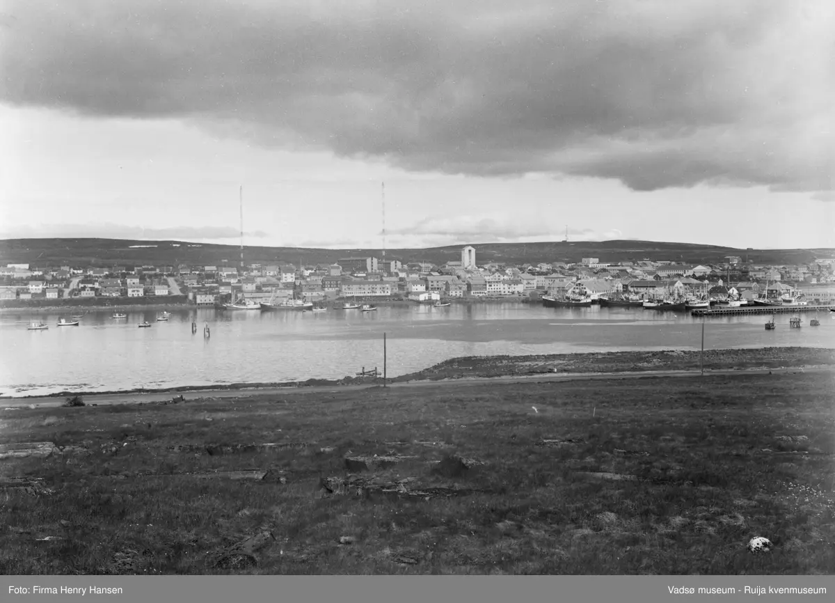Motivet viser vestre del av Vadsø sentrum og havna sett fra Vadsøya 20.07.1968. Øverst i midten av bildet ser en de to radiomastene, Fylkesbygget, Statens hus, Prestegården og Vadsø kirke. Helt til høyre i bildet sees dampskipskaia. På havna og ved kaianleggene sees store og små fiskebåter.  I forgrunnen ser man en del av veien som går fra brua til Sildoljefabrikken på Vadsøya.