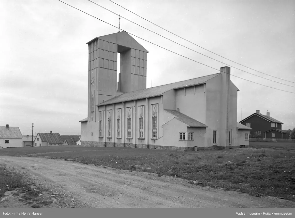 Vadsø kirke fotografert 27.07.1961. Bildet er tatt fra nord mot sydvest og vi ser nordsiden av kirken og kirketårnet samt østsiden av kirken. Nærmest i bildet ser vi deler av Aug. Trastisgate og lengst bort i høyre billedkant bebyggelse langs Amtmannsgata og Kirkegata . Til høyre for kirken ligger Vadsø Prestegård.