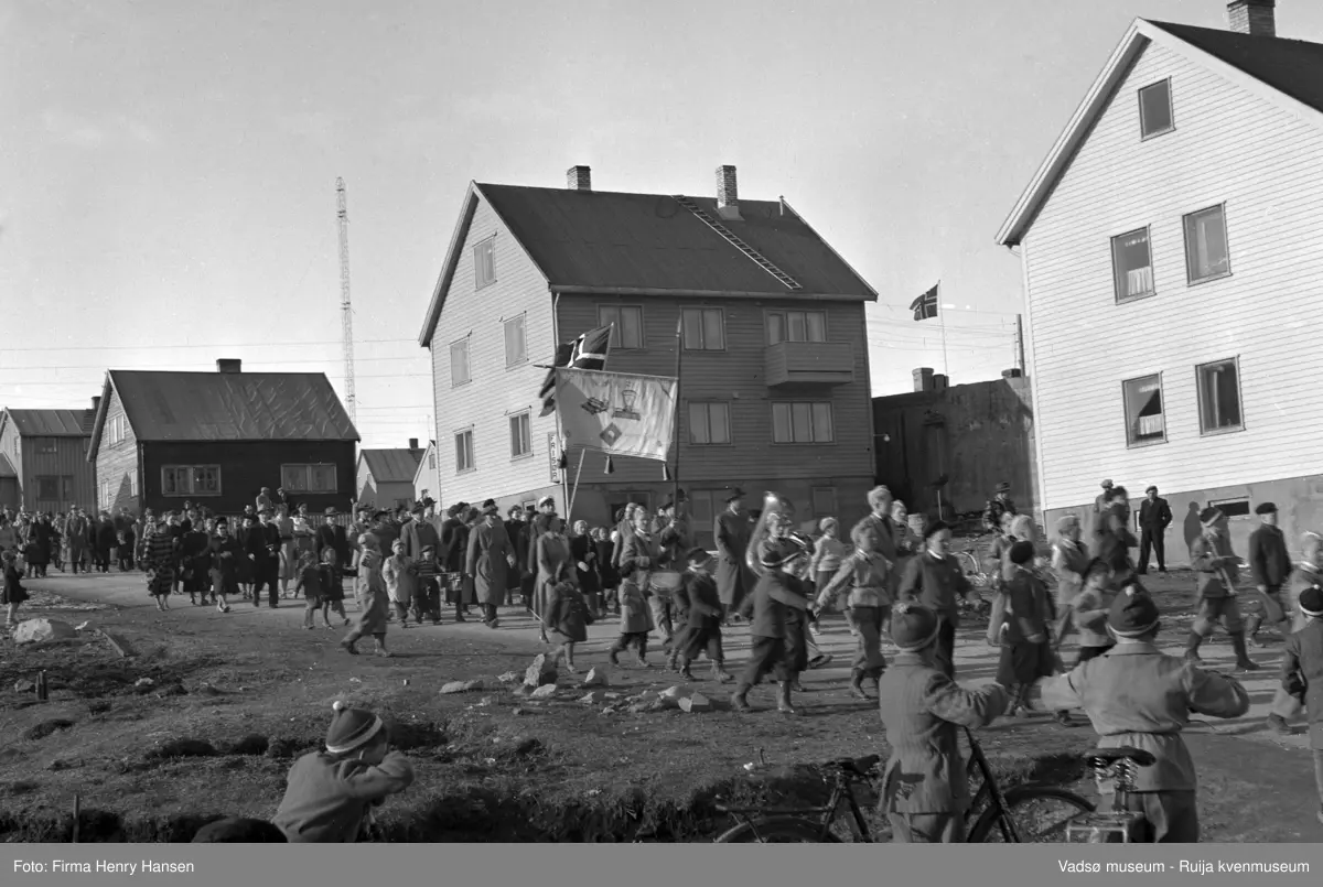 Vadsø sentrum, Kirkegata. Bildet er tatt fra syd mot nordøst. Vi ser mennesker som går i tog. De har akkurat passert krysset Oscarsgata og Kirkegata. Først i toget masjerer et musikkkorps, unge gutter delvis i uniform og med instrumenter. Rett bak masjerer voksne, noen holder en fane hvor det er bl.a. er inntegnet en butikkvekt. Langs toget ser vi voksne og barn, noen med flagg, sykler. På bygningen midt i bildet ser vi et skilt med teksten "Frisør". Her drev Beldo sin Herrefrisørforretning. En av radiomastene skimtes mot nord.