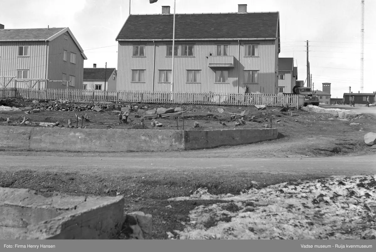 Vadsøbilder 1952 Alrik Basmas bygg. Bildet er tatt fra syd mot nord. I forkant av bildet Hvistendalsgate med deler av en grunnmur syd for veien og deler av en mur med forskaling rett nord for veien. Midt i bildet et stort gjenreisningshus i to etasjer med skifertak. I andre etasje en luftebalkong. Til høyre for bygningen står en lastbil parkert.  Innafor gjerdet som går rundt bygningen en flaggstang og tørkestativ. Til venstre innafor gjerdet ser vi deler av et annet gjenreisningshus også i to etasjer med skifertak. Til høyre i bildet går Skolegata opp mot brannstasjonen, og vi ser gjenreisningshus langs Skolegata. Øverst i bildet helt til høyre skimtes den ene av radiomastene.