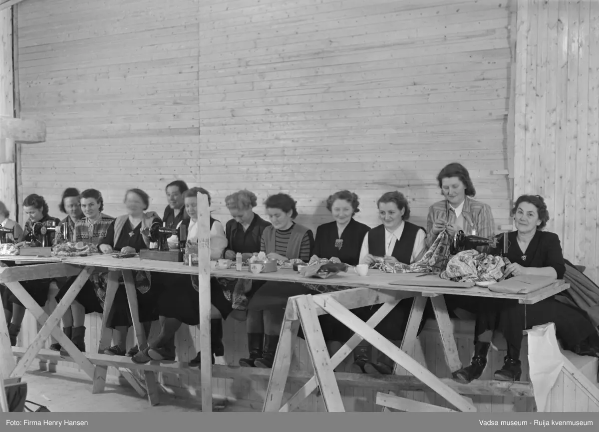 Vårbrudd 1949. Bildet er tatt inne i bygningen. Vi ser 13 flotte damer som sitter langs et provisorisk arbeidsbord og syr, både for hand og med symaskiner. Tøy, kaffekopper og matbokser ses på arbeidsbordet.
