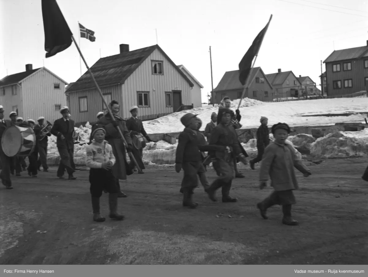 Vadsø 1.mai 1952. Folk, kvinner, barn og menn, går i tog. Noen bærer musikkinstrumenter. I forgrunnen flere barn, og to kvinner bærer flagg, det er kanskje røde flagg.  I bakgrunnen flere bolighus, flaggstang med norsk flagg ved ett av husene. Det er snø på bakken.
