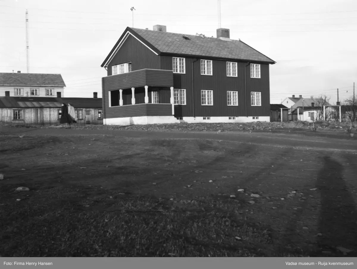 Vadsø prestegård. Bildet er tatt mot nord. Midt i bildet ser vi Vadsø prestegård, Vadsø prestebolig. Til venstre for prestegården står en langbrakke. Rett bak brakka en to-etasjes bygning med skifertak, muligens Filadelfia, og bak den ser vi deler av en rasiomast. Vi ser to kvinner spaserer på veien vest for prestegården.