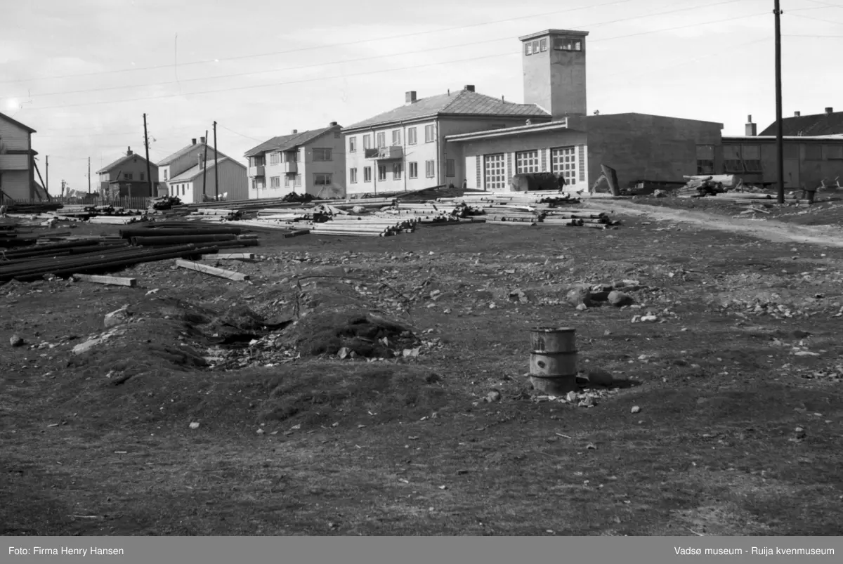 Vadsø. Bildet er tatt mot nordvest. Motivet er først og fremst brannstasjonen som ble bygd 1947-50, Vest for brannstasjonen er det to toetasjens bygninger, begge er gjenreisningshus. I forgrunnen, på en åpen plass foran brannstasjonen, ligger stolper og materialer. Et oljefat står helt foran på plassen.