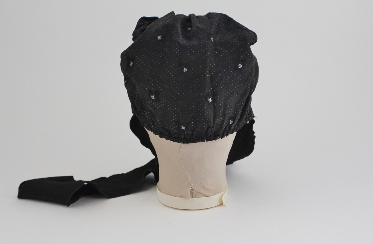 Kyse, også kalt pollelue. I mønstervevet silke i satengbinding. Stoffet er dekorert med små grå prikker med påsydde detaljer som tre små blader, rundt disse er det brodert med sort tråd ytterligere tre blader som omkranser de mindre blader. Rynket bak. Påsydd brem foran, med håndsydde rynker. Påsydde knytebånd. For i hvit bomull.